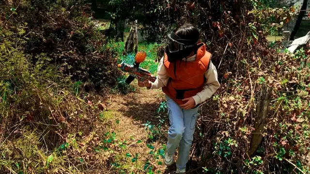 Niña jugando gelblaster con chaleco y máscara en un sendero entre arbustos del campo de Quito Woodsball